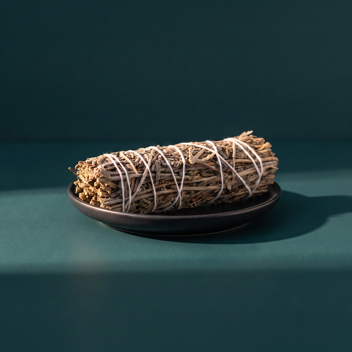 Bundle of dried lavender stems bound with white string, resting on a dark bronze dish against a deep teal background.
