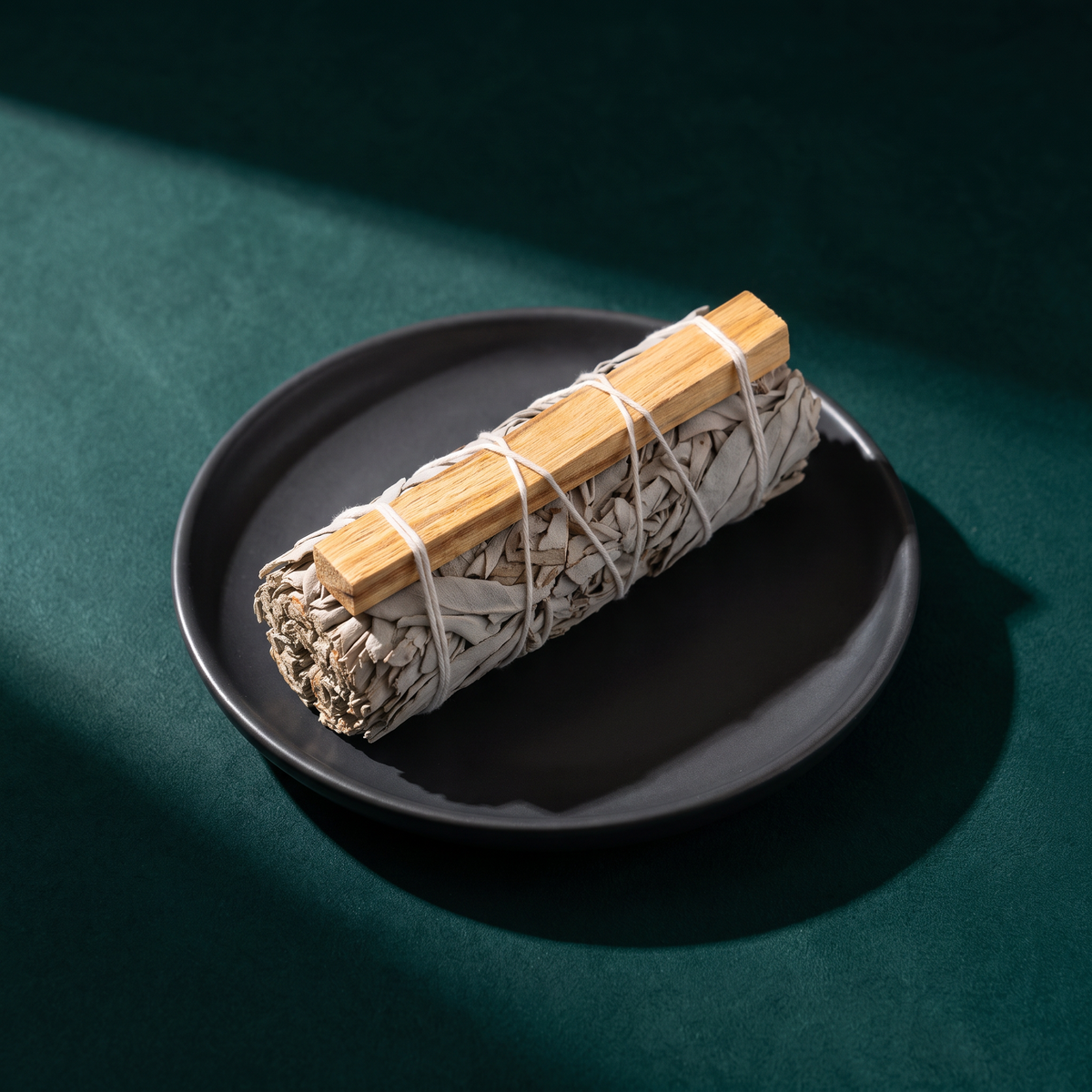 White sage bundle with a pale palo santo stick tied along its side by white string, lying on a black ceramic dish on dark green fabric in hard sunlight.