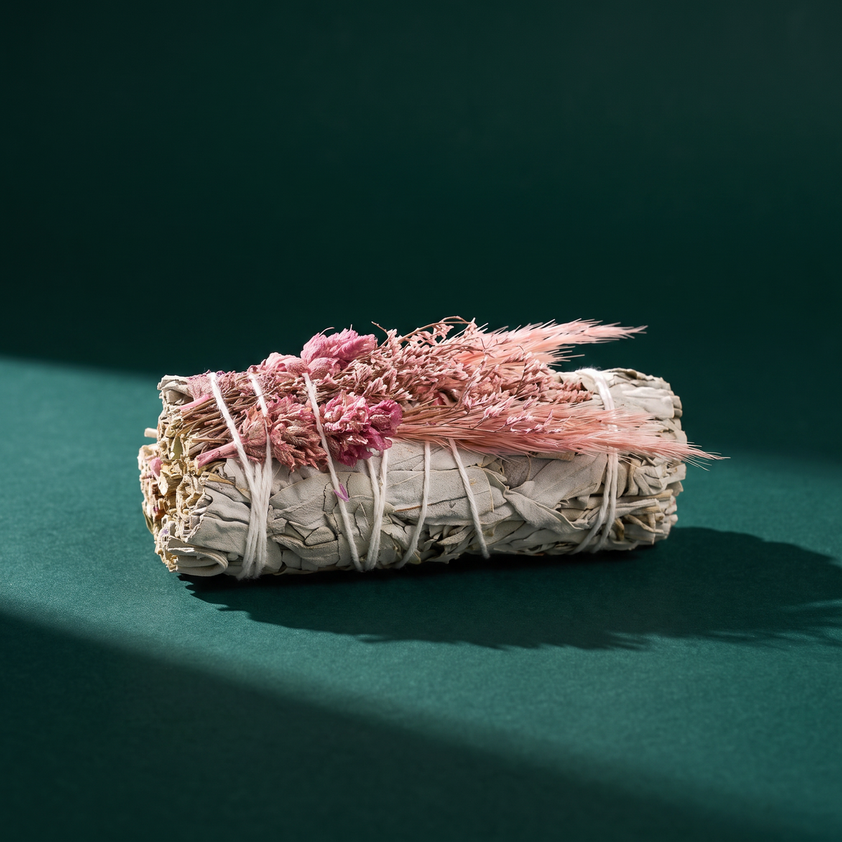 The bundle on a teal surface in raking light, seen from the side: pale gray-green sage leaves under a spray of pink statice and pink feathered grass.