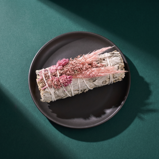 A single sage bundle on a dark plum ceramic plate against a teal background, wrapped with pink dried flowers and feathery grasses and bound in white cotton thread.