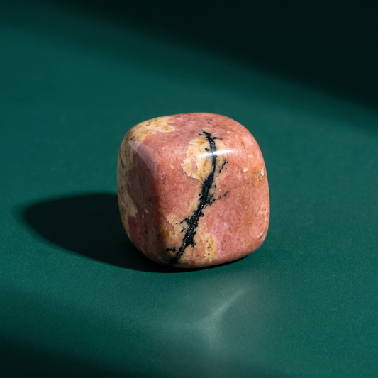 A tumbled rhodonite pocket stone in soft pink with black veining, on a dark green surface