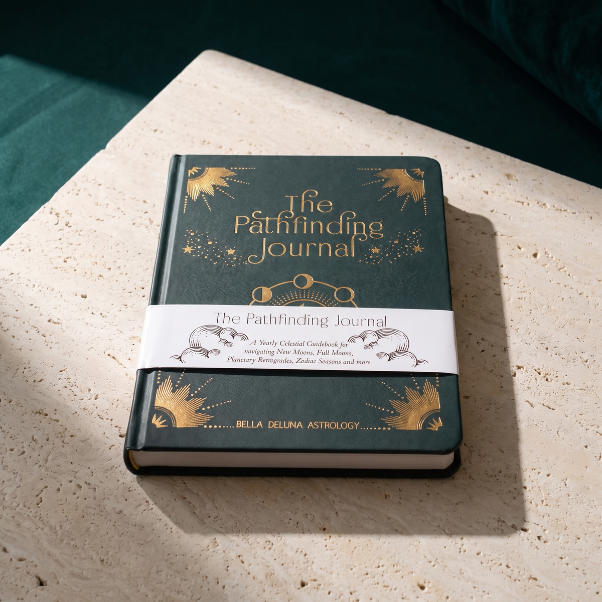 The Pathfinding Journal standing on a pale stone ledge, dark green hardcover with gold celestial artwork and its printed belly band