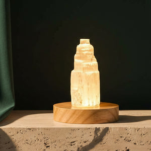 Selenite cleansing lamp lit on a light wood base, standing on a stone plinth against a dark green wall
