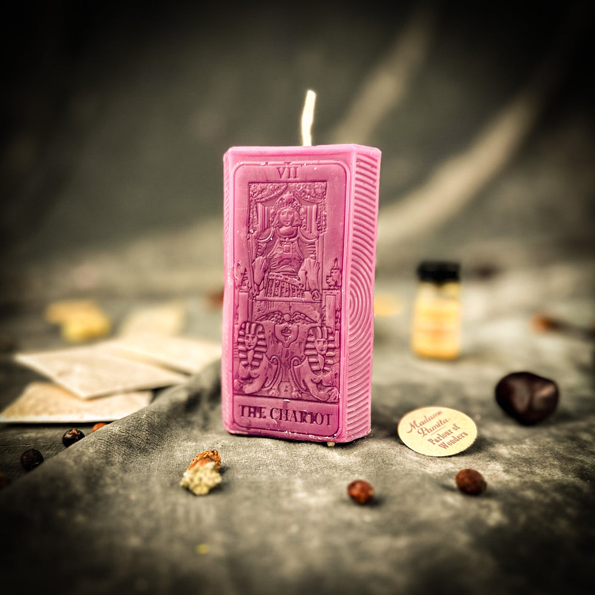 Pink beeswax The Chariot tarot card candle standing on gray cloth beside a vial of dressing oil and scattered herbs