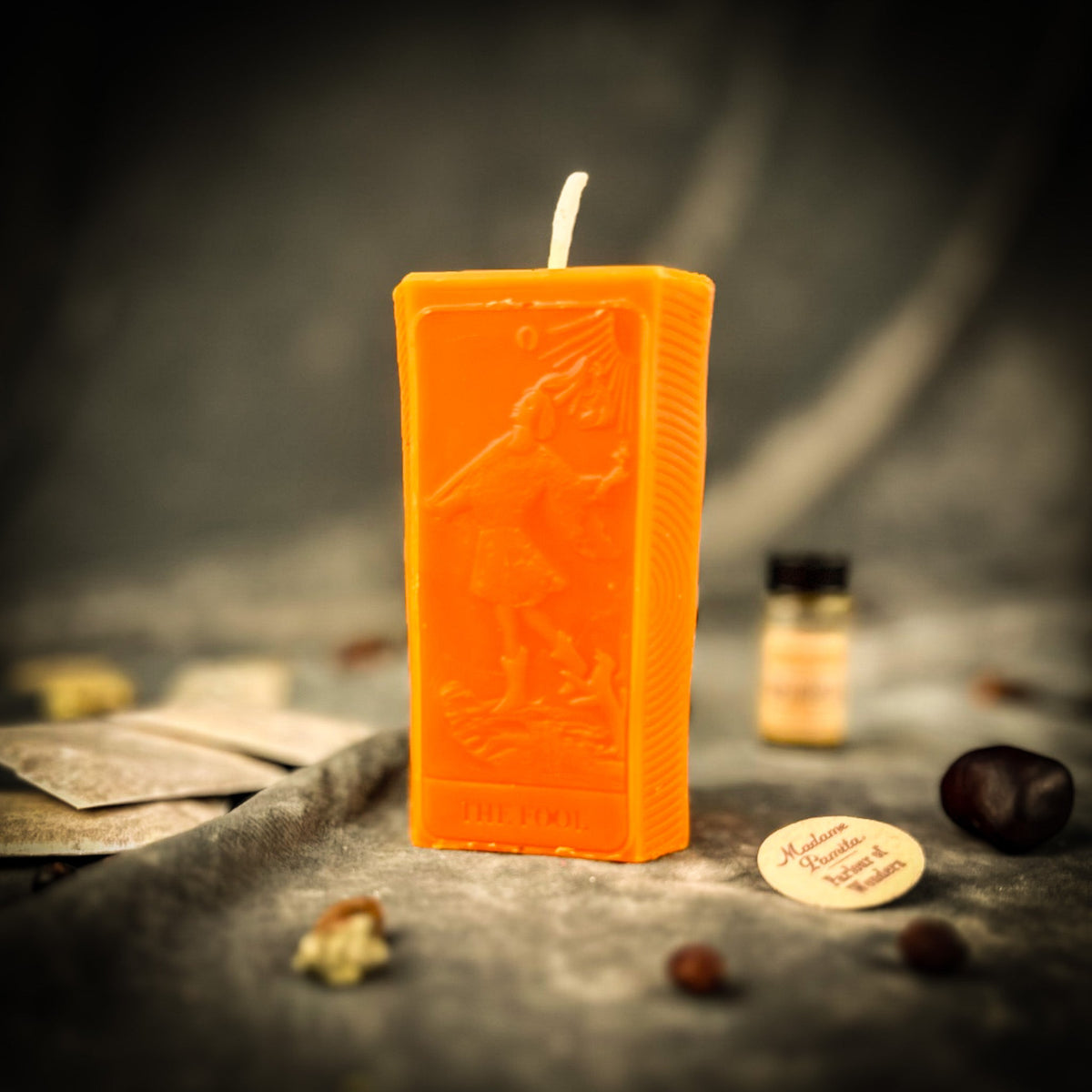 The Fool beeswax tarot candle in orange with the kit oil bottle on draped gray fabric