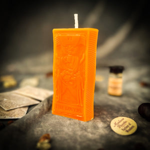 Bright orange beeswax The Lovers tarot card candle on gray cloth beside a vial of dressing oil