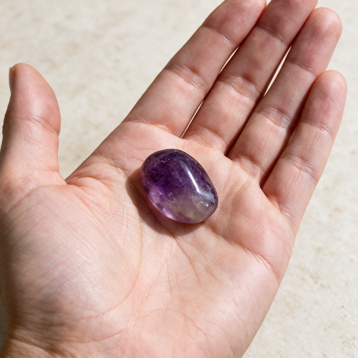 An open palm holds a polished amethyst pocket stone, purple at one end and shading to clear at the other; the stone covers about a third of the palm's width.