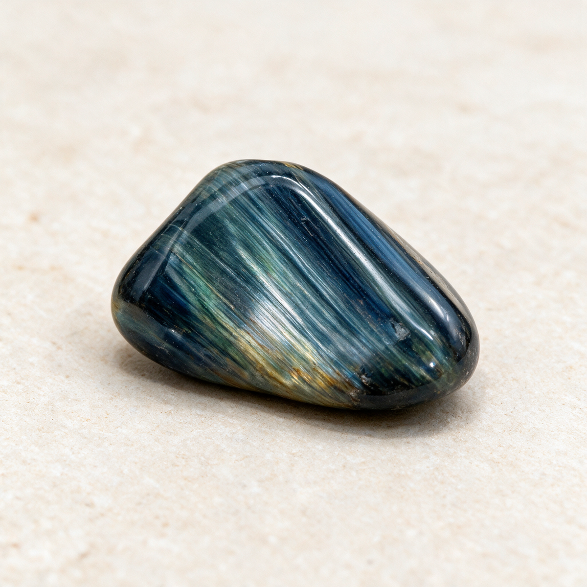 Blue tiger eye pocket stone on a warm sand-colored surface, the silky fibrous banding catching the light across its face