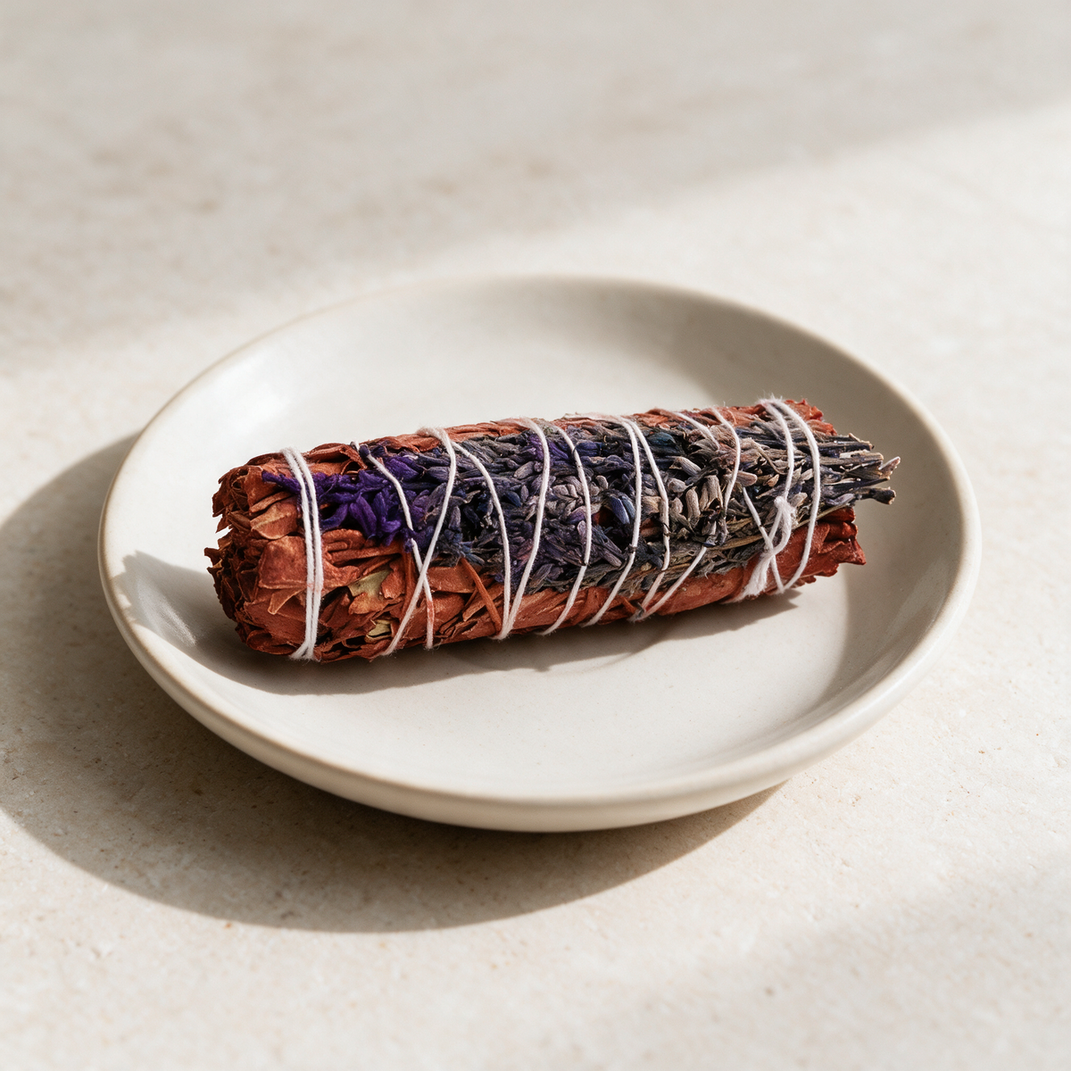 Smudge bundle of rust-red dragon's blood sage and purple lavender flowers, bound with white string, lying on a cream ceramic dish on a pale stone surface.