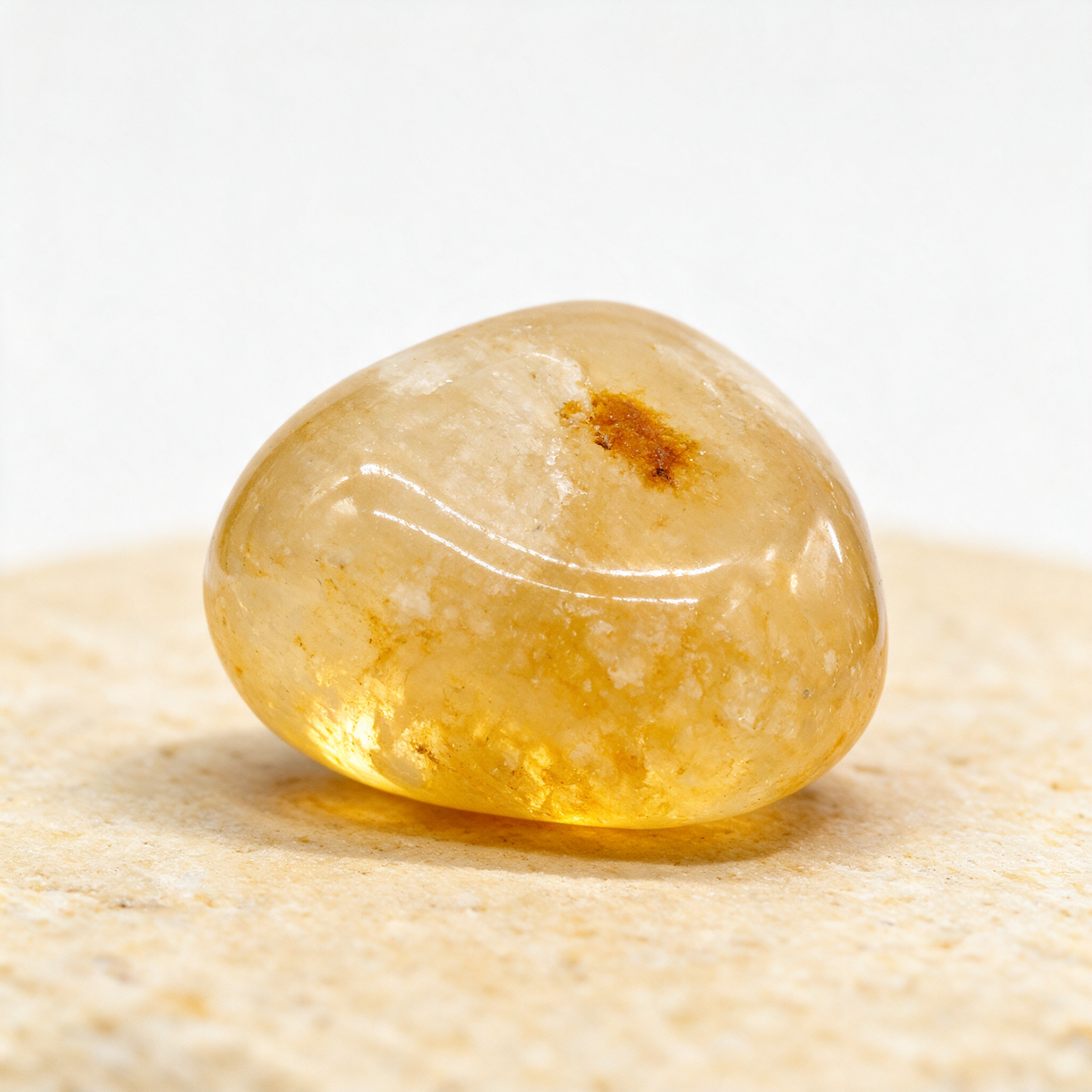 Golden healer quartz tumbled stone on pale sand, a rust-colored iron oxide inclusion visible inside
