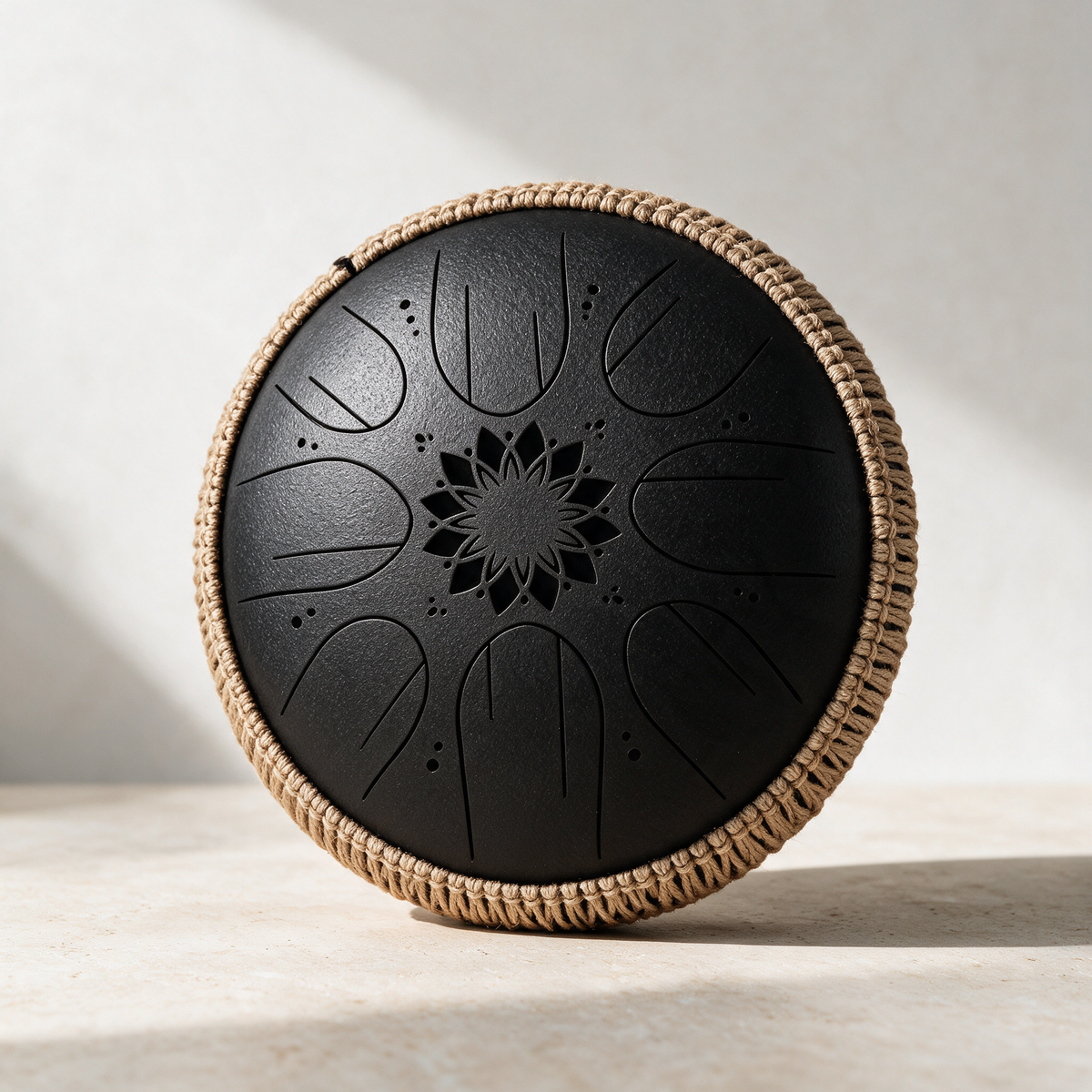 Black steel tongue drum with a rope-wrapped rim standing on a pale plaster surface in daylight