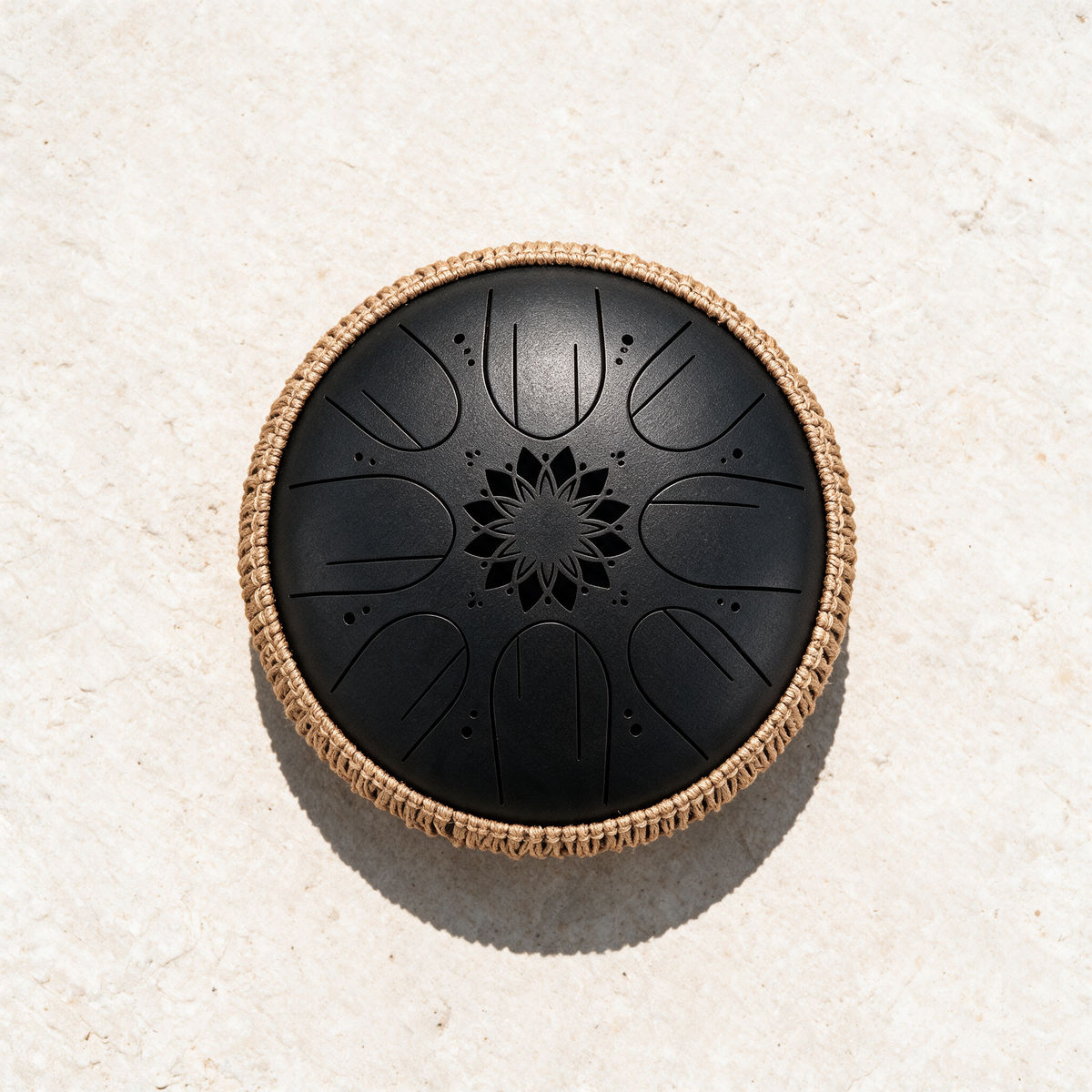 Black steel tongue drum with a rope-wrapped rim seen from above on a pale plaster surface, the eight tongues arranged around a cut lotus center