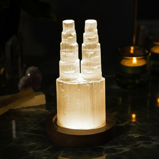 Two carved selenite towers lit side by side on a dark wooden base, standing on dark marble beside a candle.