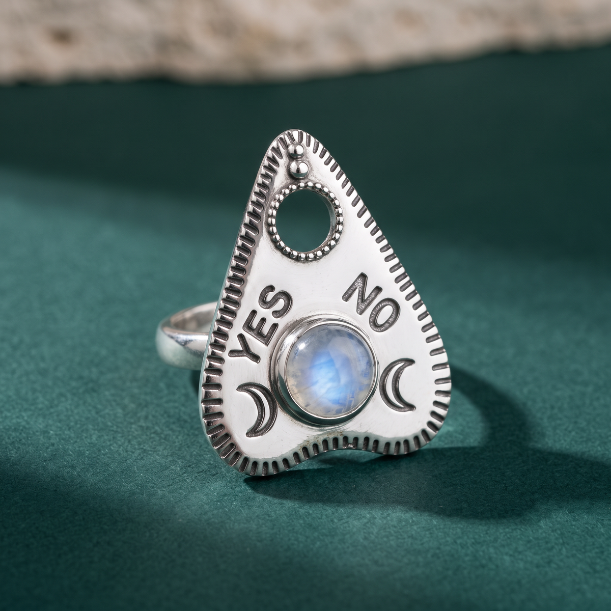 Rainbow moonstone planchette ring lying on dark green velvet beside a rough travertine edge, the YES and NO lettering catching the light