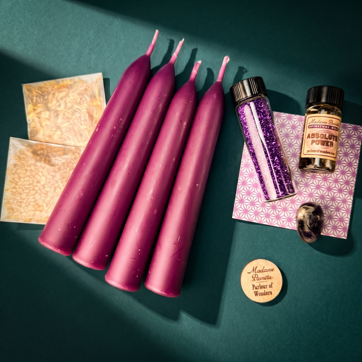 Empowerment Candle Spell Booster Pack laid out on deep green: four magenta-purple beeswax taper candles, a bottle of Absolute Power oil, a vial of purple glass glitter, two sealed herb packets, a dark tumbled stone, a petition paper and a wooden Parlour of Wonders token.
