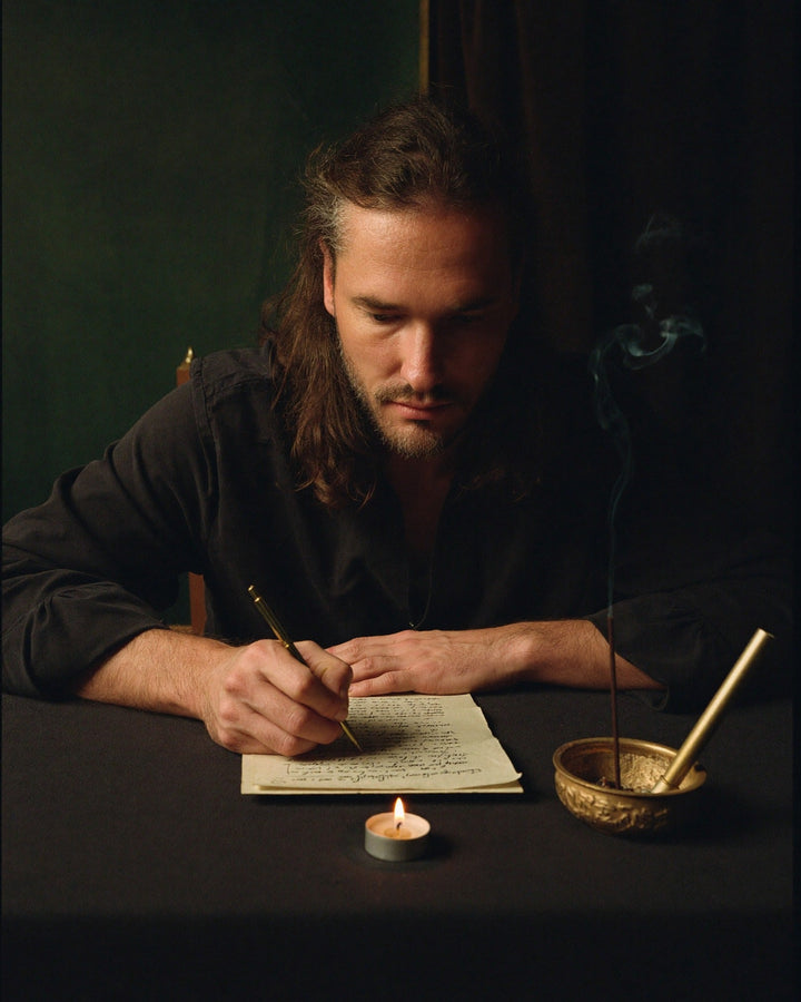 Leif Daniel writing a ritual by hand at a dark table, a tealight and smoking incense beside him