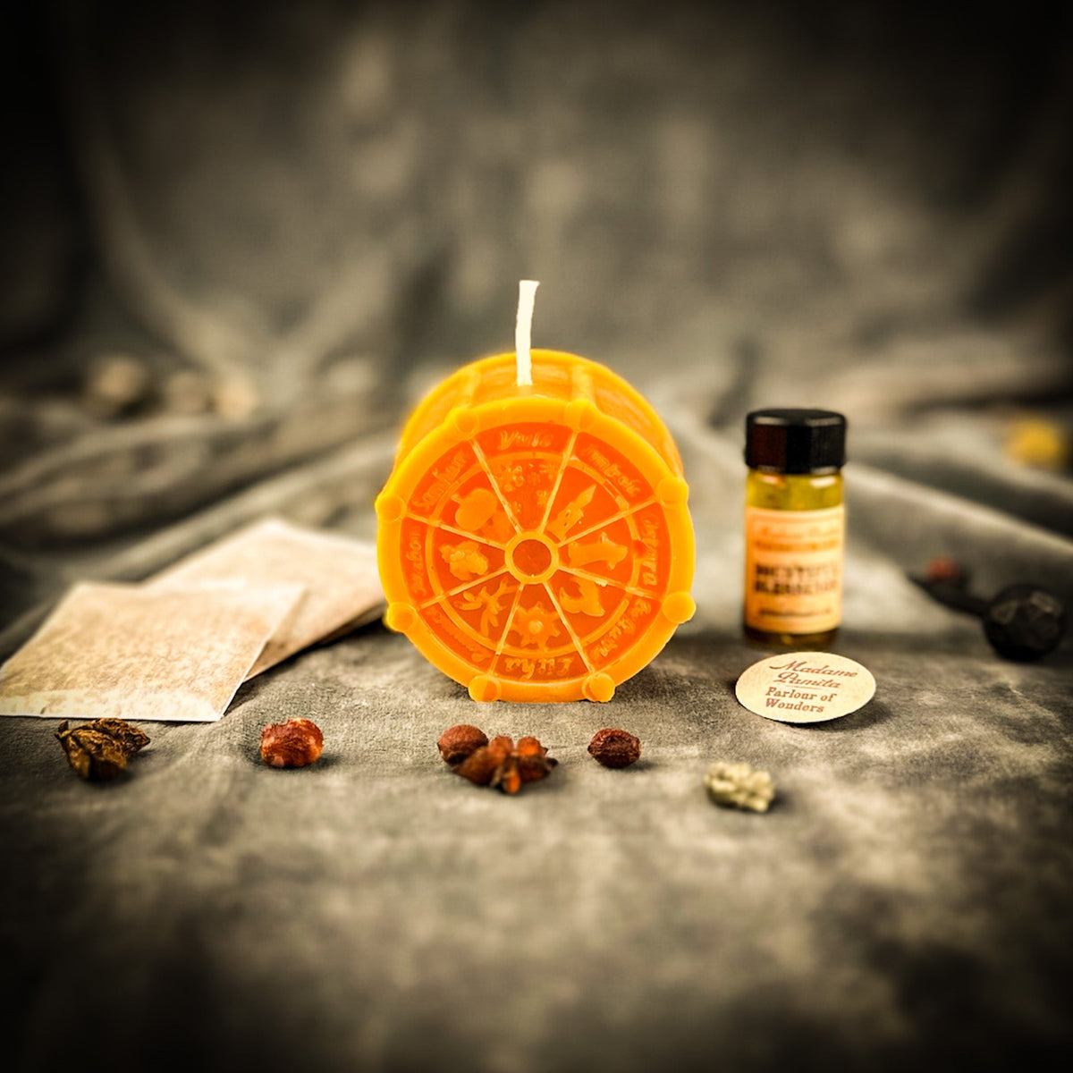 Orange Wheel of the Year candle with the oil bottle, instruction sheet and herbs behind it on gray velvet.