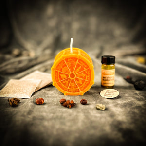 Orange Wheel of the Year candle with the oil bottle, instruction sheet and herbs behind it on gray velvet.