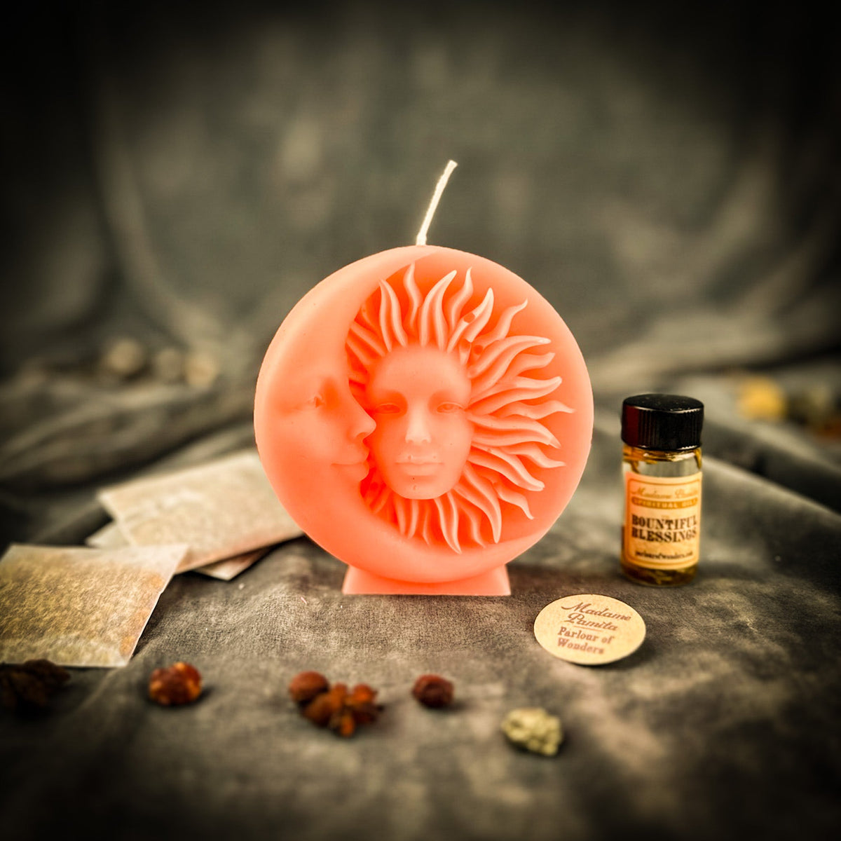 Coral sun-and-moon disc candle on gray velvet with a small vial of dressing oil, folded instruction papers and scattered berries